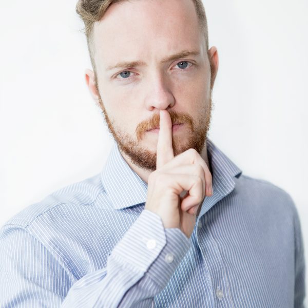 Closeup portrait of serious young man looking at camera and making silence gesture. Isolated front view on white background.